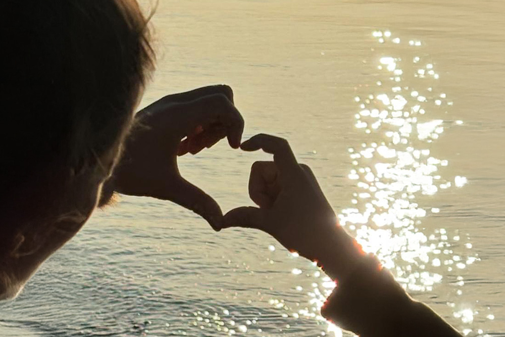 Hand einer Frau und Hand eines Kindes formen ein Herz vor dem Meer